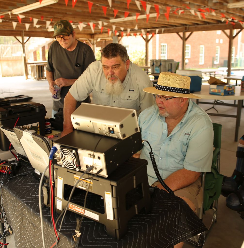Two amateur radio operators at a stack of radio equipment on a pick nick table under a pavilion on ARRL field day.