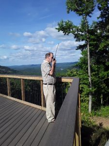 Amateur radio operator on a u0022handy talkieu0022 standing on a deck in the mountains with a mountain view behind him.