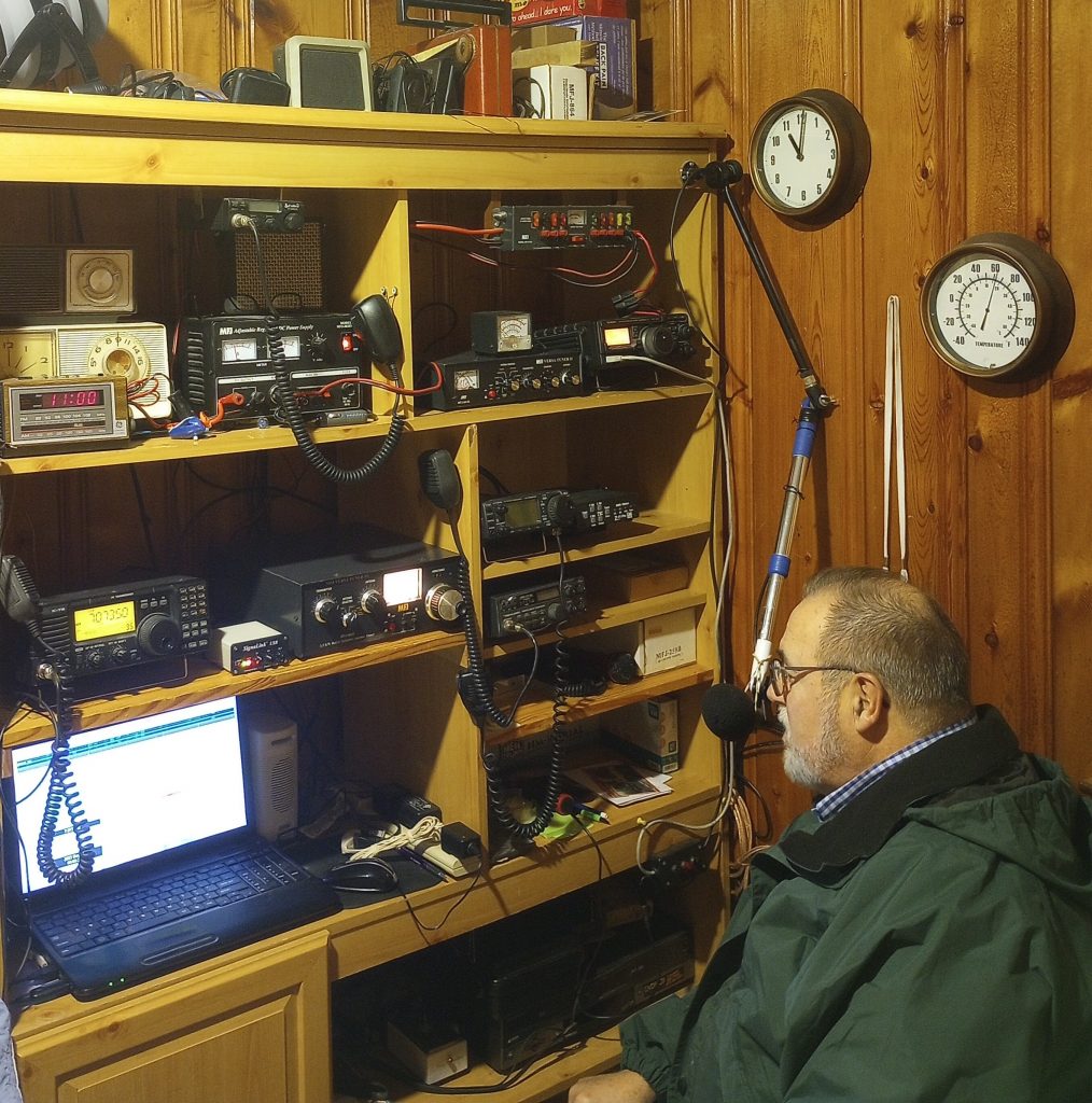 Fred Lathan sitting at a desk with shelves of radio gear, making amateur radio contacts in his home.