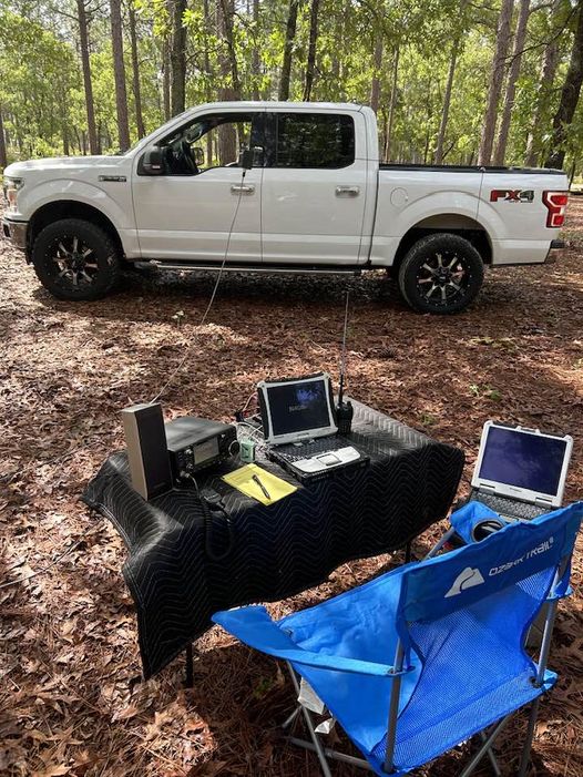 Mark Dawkins' Parks On The Air (POTA) setup. A folding chair and table with an amateur radio, computer and other radio gear in the woods.
