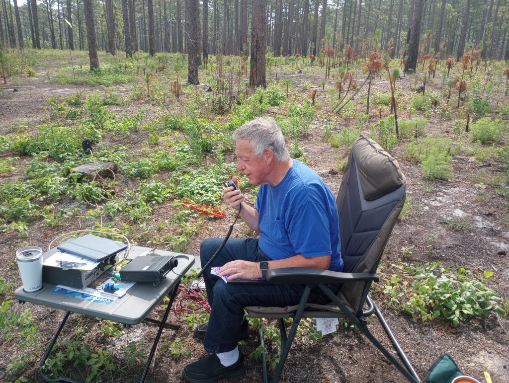 Amateur radio operator in a folding chair working a radio on a folding table in the woods, activating a park for Parks On The Air, aka POTA.