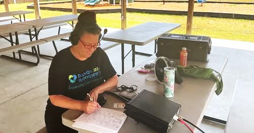 Olivia Rinehart wearing headphones and logging a contact with pencil and paper, sitting at a pick-nick table with her amateur radio setup on field day.